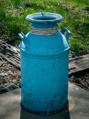 Decorative Milk Can. An aqua blue painted vintage metal milk can at the edge of a patio  backlit by the afternoon sun.