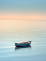 Fototapeta premium A lone wooden boat floats serenely on calm, turquoise waters under a pastel sky at sunrise, creating a peaceful and tranquil scene.