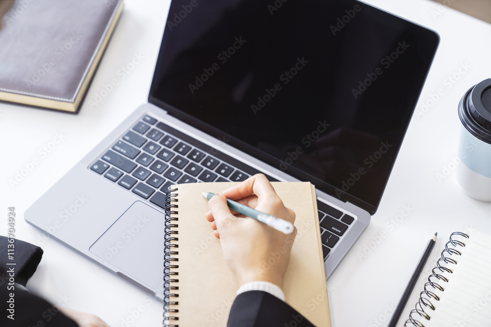 Poster detailed view of a woman's hand jotting down notes in a notepad on a modern laptop, with a blurred b