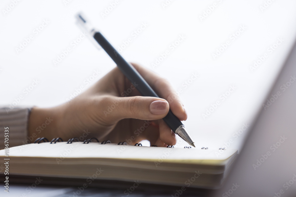 Poster close perspective of a woman's hand making entries in a notepad on a cutting-edge laptop, with a sof