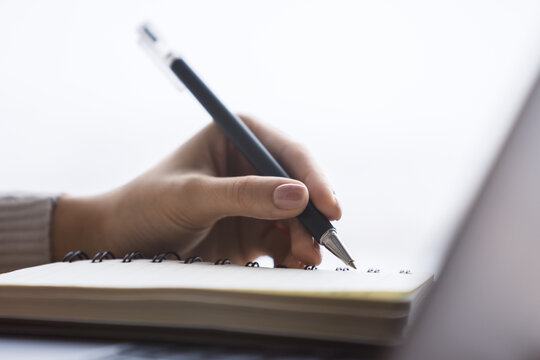 Close perspective of a woman's hand making entries in a notepad on a cutting-edge laptop, with a soft blur in the surrounding