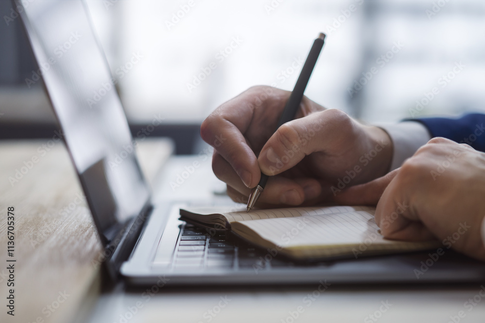 Poster Close-up capture of a man's hand as he writes in a notepad on a sleek laptop, with an out-of-focus background