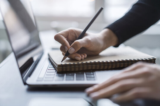 Detailed view of a woman's hand taking notes in a notepad on a cutting-edge laptop, against a blurred backdrop