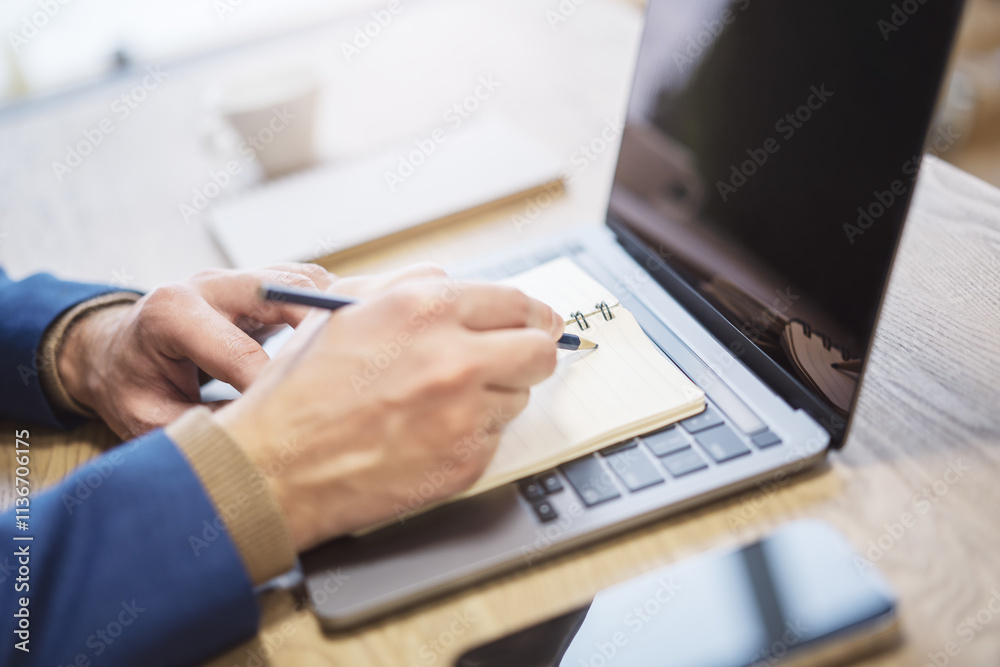 Poster Close perspective of a man's hand making entries in a notepad on a cutting-edge laptop, with a soft blur in the surrounding
