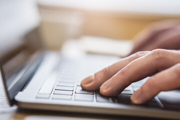 The picture shows a close-up of male hands typing on a contemporary laptop's keyboard, with a fuzzy office scene in the backdrop