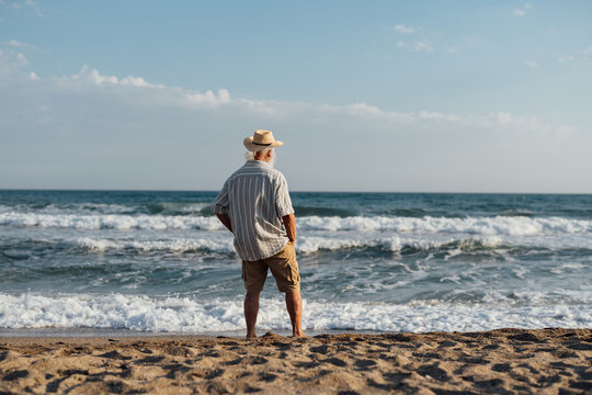 A senior man standing thoughtfully on the beach, gazing at the vast ocean waves crashing against the shore under a serene, blue sky