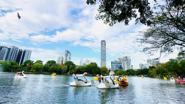 A vibrant city park scene with a tranquil lake surrounded by lush green trees and modern skyscrapers as a backdrop. In the foreground, two people in life jackets are playing in a duck boat a swan boat