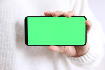 Mobile phone with green screen. Female hands holding smartphone with green screen, close-up. Girl in white sweater with phone on white background. Unrecognizable hand holding chromakey mobile phone