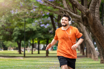 Hombre entrenando, trotando y sonriendo en un parque urbano en un hermoso día soleado. Hombre feliz corriendo mientras escucha música