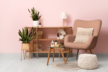 Armchair, wooden shelving unit and pouf with houseplants near pink wall in interior of living room