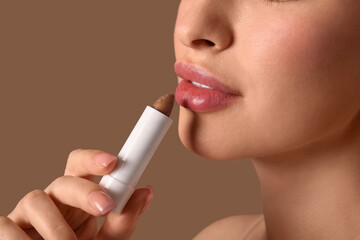 Young woman with lip balm on brown background, closeup