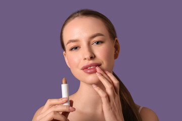 Beautiful young woman applying lip balm on purple background