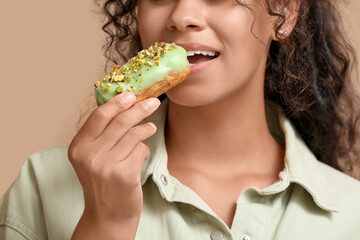 African-American woman eating tasty eclair on beige background, closeup