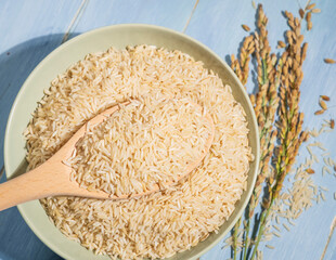 A bowl of brown rice is placed on a blue table next to a spoonful and ears of rice.The raw rice is...