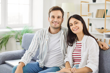 Portrait of young happy married couple sitting on sofa in living room at home. Smiling family man and woman on couch looking cheerful at camera. Harmonic relationships and Valentines day concept.