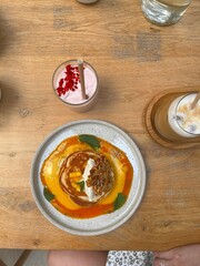 A delicious breakfast scene featuring pancakes topped with whipped cream, fresh mango, and granola on a wooden table. Accompanied by two refreshing drinks, a pink smoothie and an iced latte.