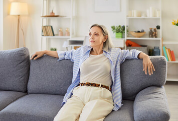Obraz premium Portrait of a happy senior woman lying comfortably on sofa at home living room, relaxing and enjoying a moment of rest. Joyful and peaceful life of a pensioner in cozy domestic living space.