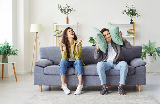 Annoyed by the noise of their neighbors, a couple sits on the sofa at home. The man covers his ears with a pillow, the woman is indignant. Concept neighbours noise, noise reduction
