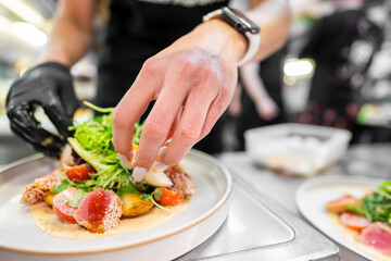 A chef meticulously plating a dish in a busy kitchen. Fresh ingredients like greens and tomatoes are arranged beautifully on a white plate, showcasing culinary artistry and attention to detail.