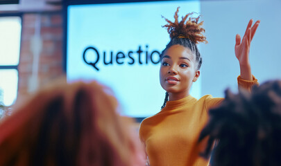 Image shows small group discussion in modern indoor space. Focus on participant with dreadlocks, raising hand for question. Screen in background reads "Questions". Teamwork, discussion and brainstorm
