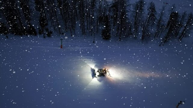 Aerial view of working snowcat on snowy alpine ski slope at winter night in snowfall. Kranjska Gora, Slovenia. Top drone view of snowcat grooming snow, ski resort, lights at dusk. Snowplough machine