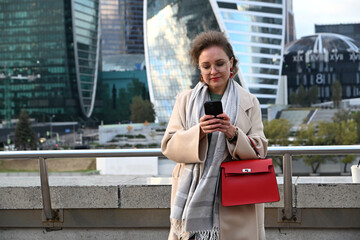 A young woman of European appearance writes a message in a messenger on a smartphone against the backdrop of Moscow City skyscrapers