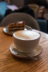 Side view of porcelain cup of pumpkin latte made from espresso, milk, ground pumpkin seeds and ground nutmeg standing on wooden table by chocolate cake. Soft focus. Copy space. Coffee break theme.