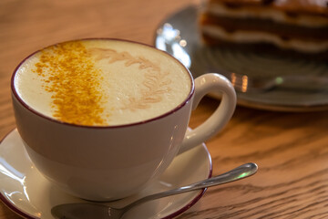 Side view of porcelain cup of pumpkin latte made from espresso, milk, ground pumpkin seeds and ground nutmeg standing on wooden table by chocolate cake. Soft focus. Copy space. Coffee break theme.