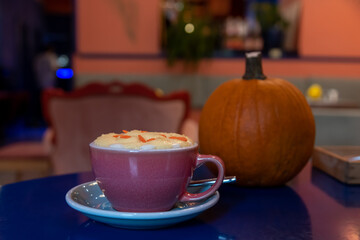 Close-up view of purple porcelain cup of pumpkin latte made from espresso, milk, whipped cream and caramel sprinkles standing on blue table next to orange pumpkin fruit in cafe or restaurant.
