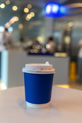 Close-up view of blue blank paper coffee cup with white plastic lid standing on round wooden table in modern cafe. Blurred espresso coffee machine in the background. Copy space. Soft focus. Hot drinks