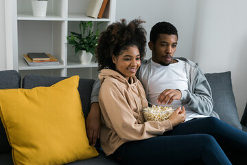 African American couple wife husband watching movie. Family pastime and leisure, watch television, rest. Relaxed man and woman watching TV series together sitting on couch at home enjoying eating - Powered by Adobe