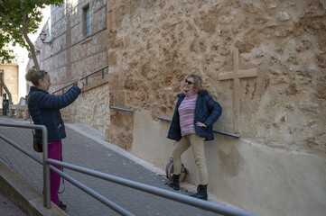 Senior woman posing playfully against a textured stone wall with a cross while her friend takes a photo in a European village