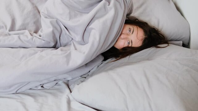 Close-up of a blue-eyed woman peeking out from under a gray blanket on a bed, playfully hiding back under the covers like a child in the morning.