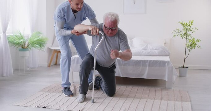 Caring young male caregiver running over to help elderly man to get up after falling in rehabilitation clinic. Friendly nurse providing assistance to pensioner patient, supporting him during walking.