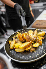 A close-up of a frying pan with golden-brown potatoes and mushrooms being sautéed. A person’s hand is visible, skillfully mixing the ingredients in a modern kitchen setting.