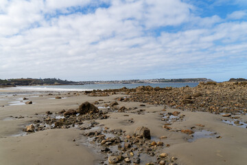 À marée basse, des cailloux parsèment le sable doré de la plage de Trez Rouz, illuminés par un doux ciel bleu parsemé de nuages blancs.