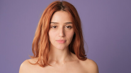 Close-up of red-haired woman looking at camera on purple background. Beautiful girl poses in studio with blank space on violet wall for your promotion