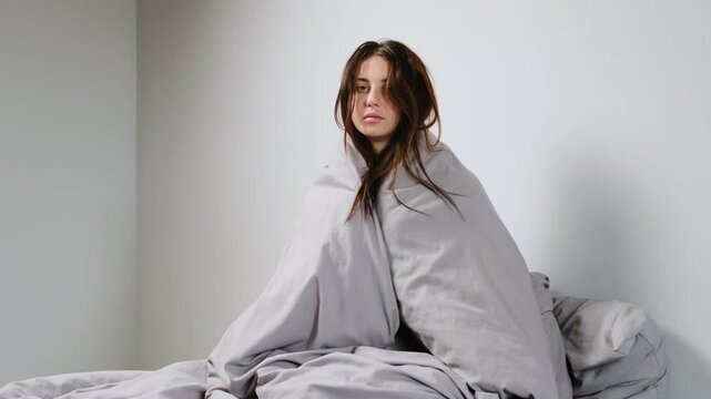 Sick young woman sitting up in bed, wrapped tightly in a gray blanket, looking pale and tired, with messy hair and a weak expression, recovering from illness in a minimal room.