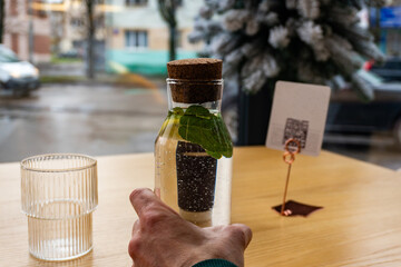 A hand is holding a carafe with water and mint leaves, placed next to a small glass, while rain...