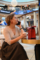 A young woman of European appearance is talking on the phone over a glass of coffee at a table in a shopping center cafe