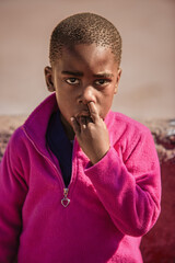 village african boy child with a finger in the nose and another in the mouth, standing in the yard at his home