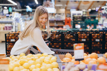 Woman choosing fresh fruits and vegetables in supermarket using smartphone
