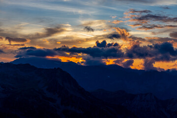 Fototapeta premium Sunset in the mountains. Red light on the sky with some clouds above the stunning mountain peaks and snow summits of the Alps. Beautiful burning skies horizon at dawn