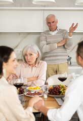 Happy young couple looking into each other's eyes sitting at table while senior parents looking at them discontentedly in the kitchen