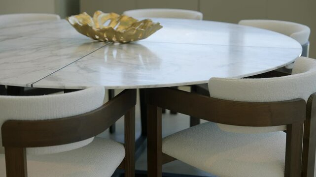 Round marble table with decorative golden bowl placed at center. Surrounding table are several modern chairs with wooden frames and white cushioned seats. Dining area