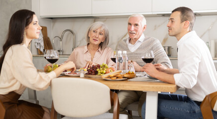 Two cheerful senior adults, man and woman, engaging in lively conversation with young couple at dinner table with wine and fruits in cozy home kitchen
