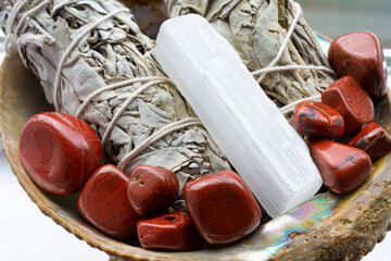 A close up image of two white sage smudge sticks in an abalone shell with red jasper crystals and selenite wand. 