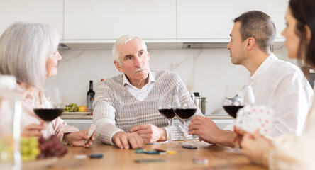 Merry old man and woman and young couple engaged in poker while having red wine in comfort of their house