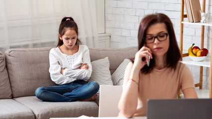 Business mom working with laptop and smartphone while her upset daughter sitting on sofa at home