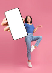 Check out this new mobile application. Excited young Asian lady jumping, shouting OMG, presenting smartphone with empty screen over pink studio background, mockup for website or app